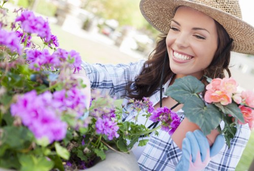 A woman with a wide-brimmed straw hat and wearing light blue gardening gloves is smiling as she tends to a variety of flowering plants in a well-maintained garden. The garden features lush green grass and a combination of vibrant purple, pink, and orange blossoms, possibly in containers or flower beds. In the background, there are out-of-focus trees and a landscaped outdoor space, indicating a sunny day with natural light enhancing the colours. The woman's focus on the plants suggests she is engaged in gardening activities such as planting or pruning, supported by her attire suitable for outdoor gardening. The scene captures a tidy, inviting garden environment with healthy, blooming flowers and neatly maintained plantings, illustrating professional gardening or horticultural care typical of services provided in the Seven Kings area, close to Ilford in east London, underlining the offerings of Hedge Trimming Seven Kings for gardening and green space maintenance.